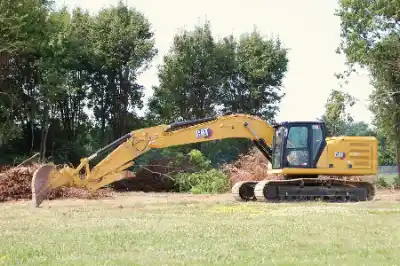 A yellow excavator sits on grass near trees and brush